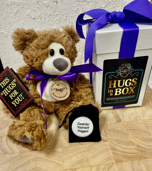 Small plush teddy bear with purple ribbon, sitting next to a white “Hugs in a Box” gift set tied with a purple bow.