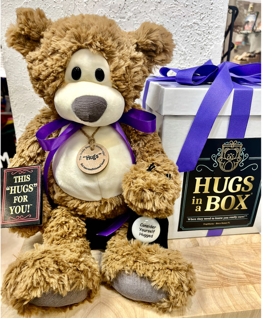 Large cuddly teddy bear featuring a purple ribbon and Hugs tag, displayed with a white Hugs in a Box gift set wrapped with a purple bow for a warm, thoughtful present.